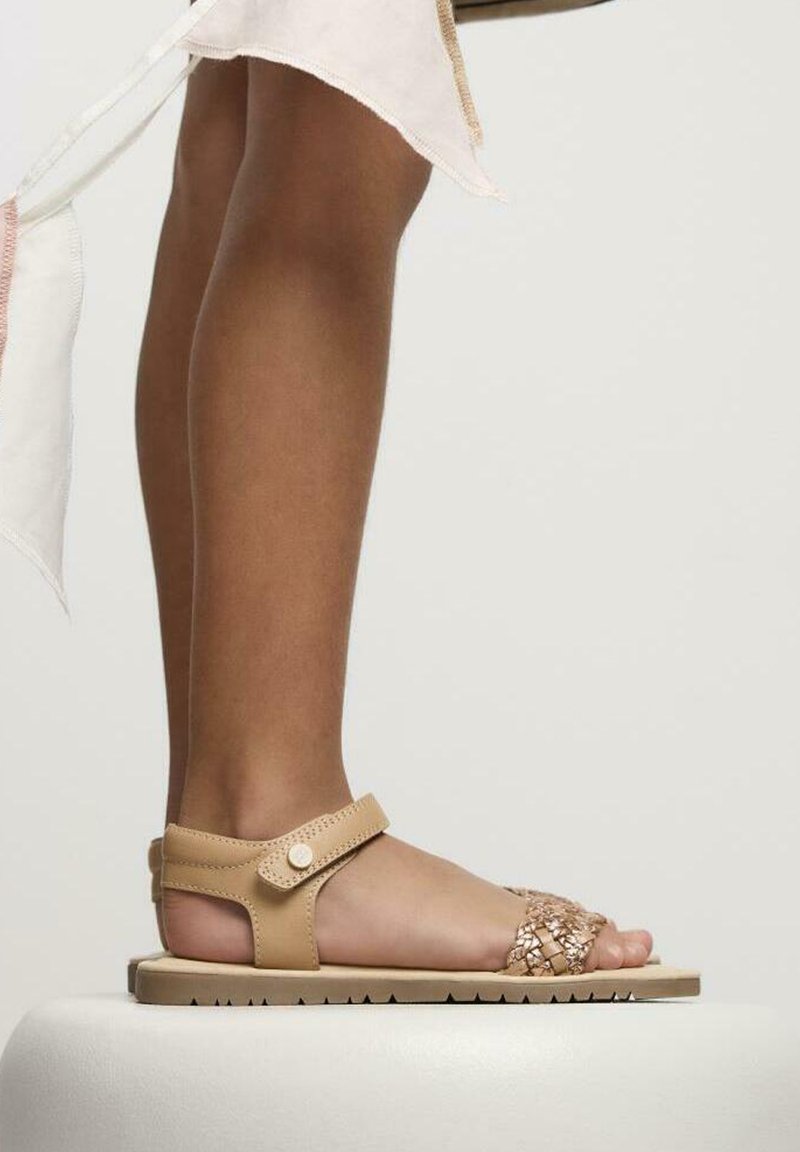 Sandal de color beige que presenta una tira trenzada, una banda ancha para el tobillo con cierre de botón a presión, y una suela texturizada. Colocada sobre una superficie blanca, enfocándose en el pie.