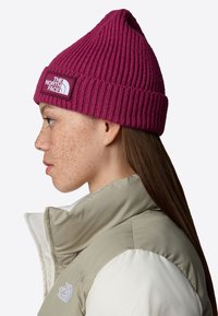 Zgniły beanie w głębokim magencie, z prążkowaną fakturą i prostokątną łatą z logo. Nosi się z beżową i białą kurtką puchową.