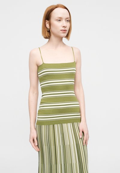 Νεαρή γυναίκα με κοντά κόκκινα μαλλιά, που φοράει χρυσούς κρίκους και πράσινο-άσπρο ριγέ αμάνικο φόρεμα, στέκεται μπροστά σε απλό φόντο.