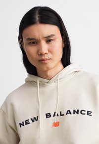 Cremfarbener Hoodie mit großem schwarzem "NEW BALANCE"-Logo und orangefarbenen Akzenten. Verfügt über Kordeln und eine weiche Textur.