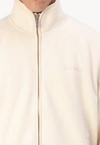 Cream-colored fleece jacket with a high collar, zip closure, and a subtle embroidered logo on the left chest. Soft texture.