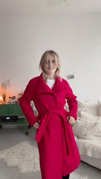 Νέα γυναίκα με κόκκινο παλτό με ζώνη στέκεται σε ένα ζεστό σαλόνι με πράσινη ντουλάπα, λευκό καναπέ και μαλακό χαλί.