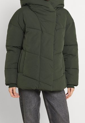 Personne portant une veste d'hiver matelassée vert foncé avec des poches avant et un jean gris sur un fond uni.