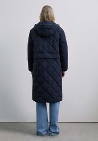Navy quilted puffer coat with a hood, featuring a belted waist and long sleeves, paired with light blue wide-leg jeans.