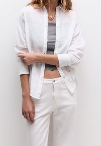 Chemise boutonnée en lin blanc portée sur un débardeur gris, accompagnée de pantalons blancs taille haute. Comprend des boutons, une coupe décontractée et des manches retroussées.