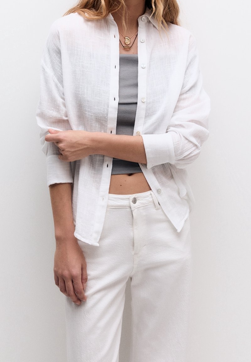 Chemise boutonnée en lin blanc portée sur un débardeur gris, accompagnée de pantalons blancs taille haute. Comprend des boutons, une coupe décontractée et des manches retroussées.