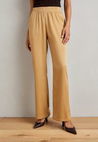 Gouden wijde broek met een gestructureerde stof, elastische tailleband en zijzakken, gecombineerd met donkerbruine hakken op een houten vloer.