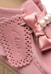 Zapatilla de ante rosa con etiqueta de cordones en forma de onda, un lazo de cintas de satén y acentos de perla, con suela texturada y un interior transpirable.