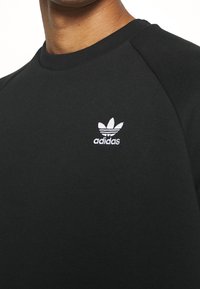 Svart bomullströja med rund halsringning och raglanärmar, med en vit broderad Adidas-logotyp på vänster bröst.