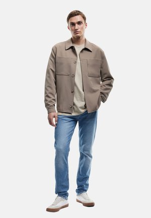 Beige knoopjack met twee borstzakken, gedragen met een lichtbeige t-shirt, blauwe jeans en witte sneakers met bruine accenten.