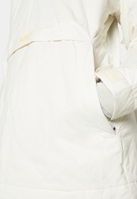 Weiße gesteppte Jacke mit verstellbarem Bündchen, einer großen Fronttasche und einem Roxy-Logo in sanftem Gelb. Der Stoff wirkt glatt mit mattem Finish.