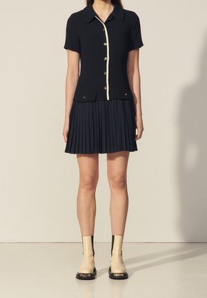 Kobieta ubrana w czarną, krótką koszulę z guzikami i białym wykończeniem, granatową plisowaną spódnicę oraz kremowe botki z czarnymi podeszwami.