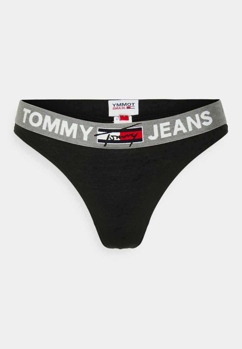 Tanga negro con una cinturilla elástica gris que presenta "TOMMY JEANS" en letras blancas y una pequeña etiqueta de logo en la parte superior.