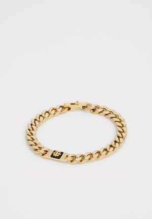 Bracelet en chaîne dorée avec des maillons épais et un emblème rectangulaire noir et or comportant un petit détail en forme de fleur, fermoir visible.