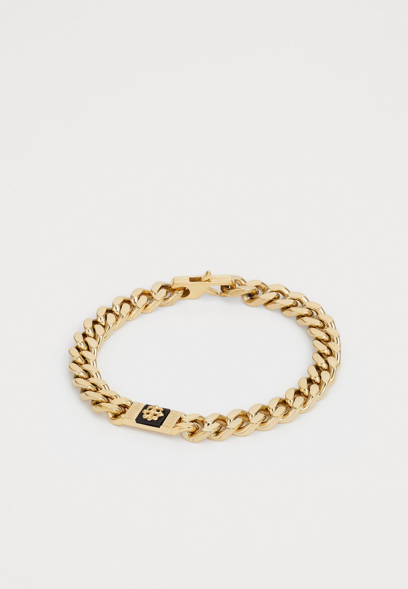 Bracelet en chaîne dorée avec des maillons épais et un emblème rectangulaire noir et or comportant un petit détail en forme de fleur, fermoir visible.