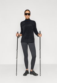 Melns, fakturēts adīts džemperis ar augstu apkakli, saskaņots ar tumšām legingām un melniem pārgājienu apaviem. Tur pelēkas pārgājienu nūjas; valkā saulesbrilles.