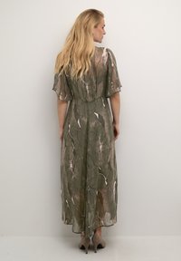 Robe longue verte à motifs avec des manches courtes et fluides, présentant un mélange de couleurs claires et foncées dans un design abstrait. Le mannequin porte des talons beiges.