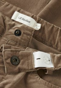 Pantaloni in corduroy marroni con vita regolabile, dotati di un bottone a scatto nero e una linguetta elastica bianca interna.