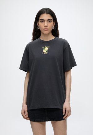 Femme aux longs cheveux foncés portant des boucles d’oreilles créoles argentées, un T-shirt noir oversize avec un imprimé pêche, et une jupe en denim noire, debout sur fond blanc.