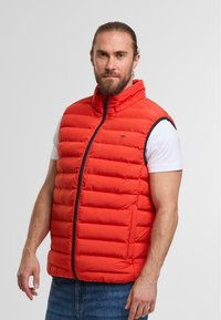 Orangen gesteppte Weste mit hohem Kragen, Reißverschluss vorne und kontrastierenden Besätzen. Über einem weißen T-Shirt getragen, kombiniert mit Jeans.