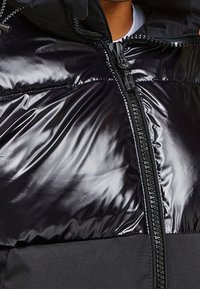 Svart puffjacka med en glänsande, slät ytteryta, dragkedjestängning och huva. Materialet har en glansig textur med en kontrasterande matt nederdel.