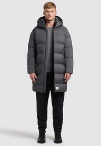Szary kurtka puchowa z kapturem, o pudełkowym kroju, pikowanym wykończeniu i subtelnym logo na dolnym wykończeniu. W zestawieniu z czarnymi spodniami i butami.