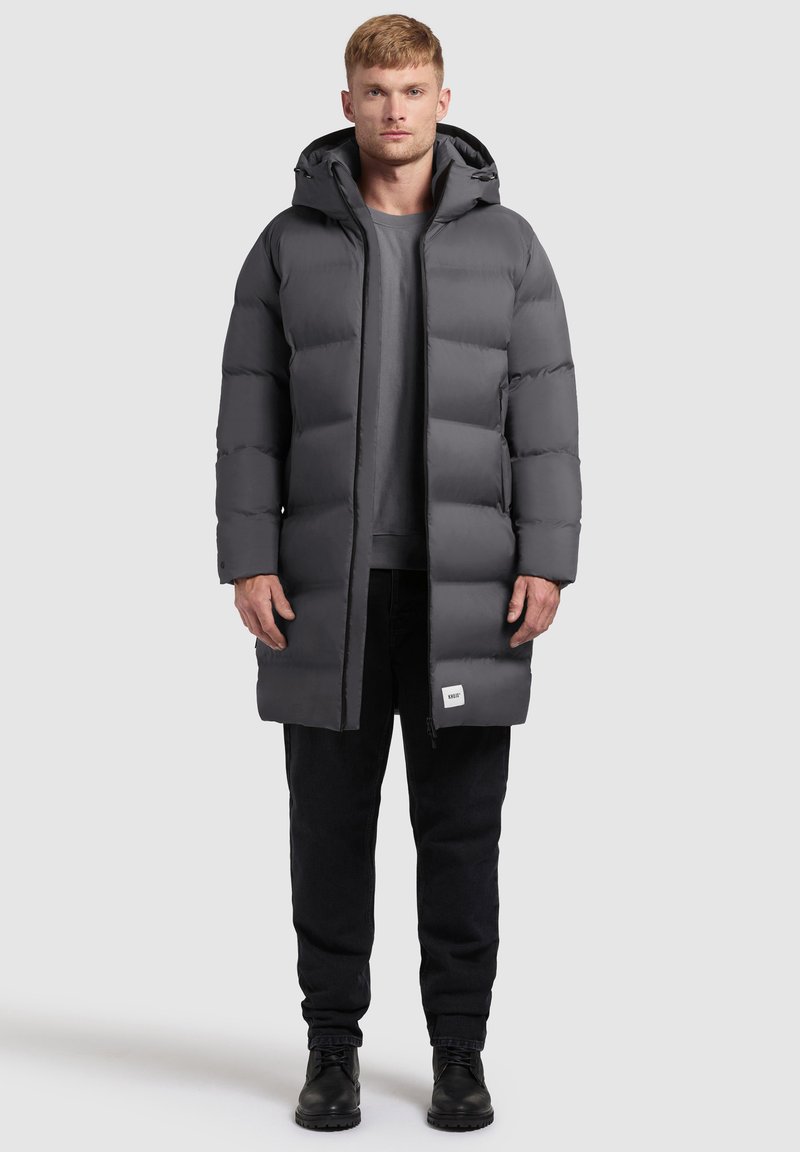 Szary kurtka puchowa z kapturem, o pudełkowym kroju, pikowanym wykończeniu i subtelnym logo na dolnym wykończeniu. W zestawieniu z czarnymi spodniami i butami.