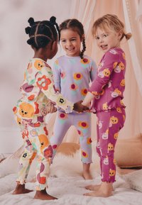 Drei Kinder in bunten Schlafanzügen: eines mit Blumenmuster, eines in Helllila mit Blumen und eines in Rosa mit Tiergesichtern.