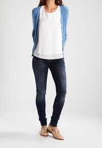 Ljusblå cardigan över en vit blus, kombinerad med mörkblå tighta jeans och beige snörskor med platt häl och dekorativ slarv.