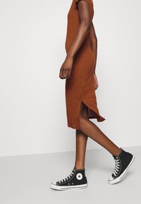 Ruskea polvipituinen mekko, jossa on lyhyet hihat ja sivuaukot, yhdistetty mustiin korkeisiin tennareihin, joissa on valkoisia yksityiskohtia ja nauhoja.