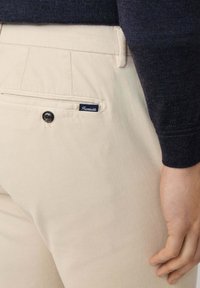 Beżowe bawełniane spodnie mają tylną kieszeń z granatową etykietą i czarnym akcentem guzikowym. Kieszeń ma czysty, minimalistyczny design.