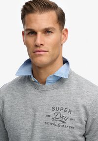 Grijze sweatshirt met een ronde hals en ribgebreide manchetten, voorzien van een zwart geprint logo. Lichtblauw overhemd met kraag zichtbaar eronder.