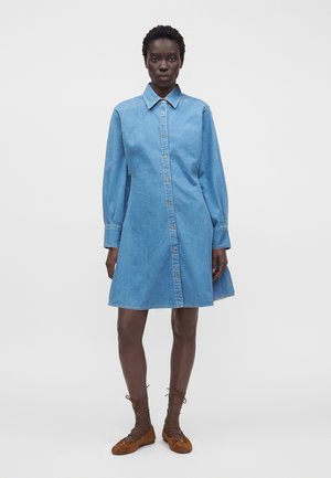 Femme debout portant une robe-chemise en jean bleu clair à manches longues avec fermeture par boutons et des chaussures plates marron à lacets.