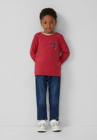 Czerwona koszula z długim rękawem z białym kołnierzykiem i małym kwiatowym wzorem na piersi, w połączeniu z niebieskimi dżinsami i jasnymi trampkami.