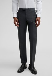 Dunkelgraue, maßgeschneiderte Hose aus strukturiertem Stoff, mit flacher Vorderseite, Gürtelschlaufen und leichten Bügelfalten, kombiniert mit schwarzen Anzugschuhen.