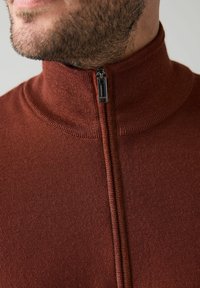 Rozsdabarna cipzáras pulóver, magas nyakkal és sima kötött textúrával. Elején fekete cipzárral.