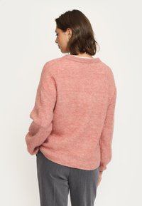 Rosa, fluffig tröja med avslappnad passform, rundad halsringning och sänkta axlar. Kombinerad med grå byxor som visar en mjuk textur.