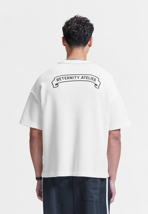 Mand med kort krøllet hår bærer en løs, hvid tekstureret kortærmet skjorte med teksten "Reternity Atelier" øverst på ryggen.