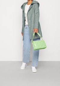 Borsa a tracolla verde trapuntata con forma strutturata, dotata di un manico corto e superficie liscia. Indossata con un cappotto grigio con cappuccio e denim chiaro.