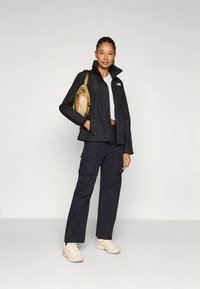 Svart vanntett jakke med høy krage, kombinert med marineblå cargobukser og kremfargede joggesko. Modellen bærer en beige skulderveske.