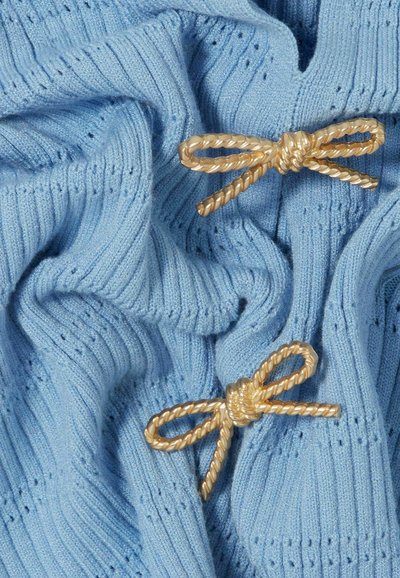 Il tessuto a maglia blu chiaro presenta una texture a costine e due fiocchi decorativi in corda dorata, che aggiungono dettaglio e interesse visivo.