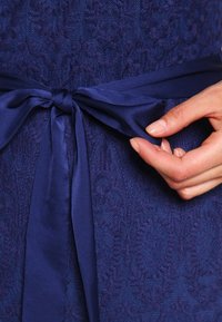 Námořnický textil s texturovaným květinovým vzorem, ozdobený saténovou stuhou uvázanou v pase, kterou drží ruka.