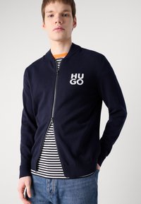 Veste zippée marine avec des poignets et un ourlet côtelés, ornée d'un logo "HUGO" blanc sur la poitrine gauche. Portée par-dessus un t-shirt rayé.