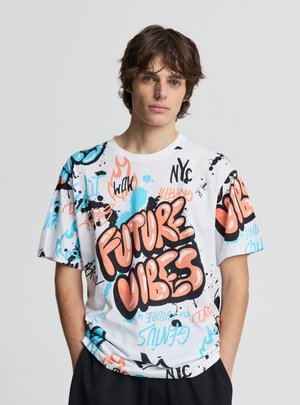 Giovane uomo con capelli castani di media lunghezza indossa una T-shirt bianca con motivo grafico con testo colorato in stile graffiti "Future Vibes" e disegni astratti.