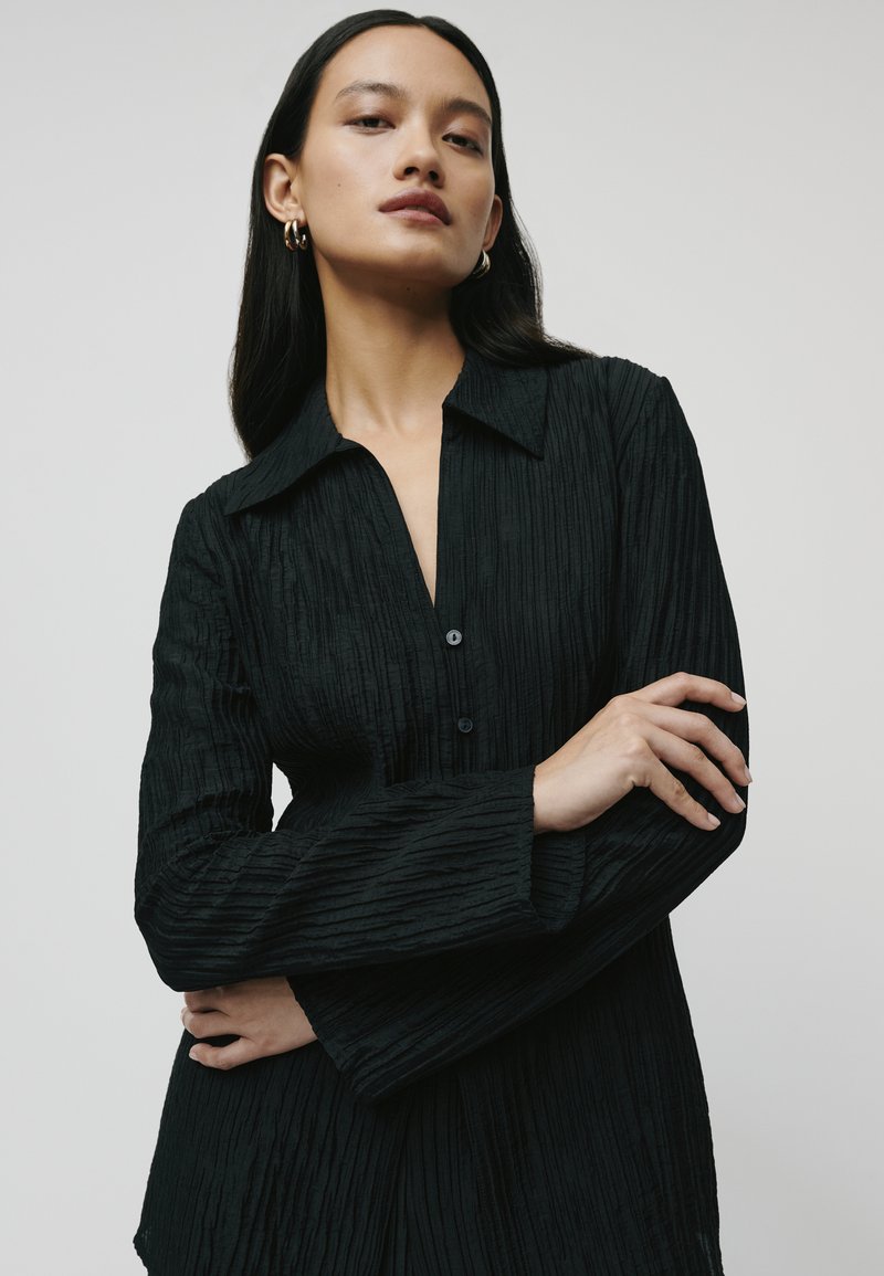 Black, textured shirt with a collared neckline, long sleeves, and button closure, featuring vertical pleats for added dimension.