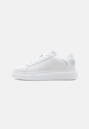 Zapatilla blanca de corte bajo con suela gruesa, parte delantera con cordones y perforaciones sutiles en la parte superior de cuero liso, mostrada en vista de perfil.