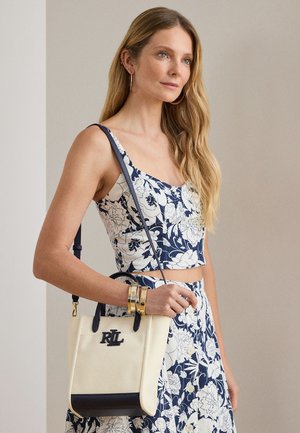 Γυναίκα με μακριά ξανθά μαλλιά που φοράει μπλε και λευκό λουλουδάτο κοντό τοπ και φούστα, κρατώντας μπεζ και μαύρη τσάντα με το λογότυπο "RLL".