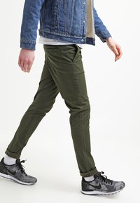 Olivgröna chinos med slim fit och sidofickor, tillsammans med en jeansjacka och svarta sneakers med en texturerad design.