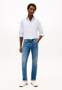 Camicia azzurra a righe con bottoni, vestibilità comoda, abbinata a jeans a gamba dritta di media lavatura e scarpe da ginnastica bianche. Abbigliamento semplice senza fantasie.