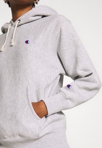 Personne portant un sweat à capuche Champion gris clair avec poche avant et logos sur la poitrine et la manche, main reposant dans la poche.